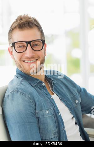 Uomo adulto seduto sorridente guardando la macchina fotografica vicino alle finestre con occhiali neri camicia in denim T-shirt bianca. Ritratto, naturale, luce del giorno, luminoso, occhiali, bea Foto Stock