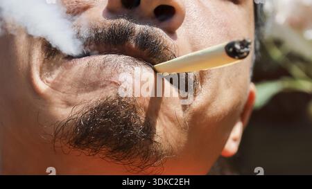 Uomo rilassato che fuma l'articolazione di cannabis. Primo piano su bocca e baffi con fumo bianco. rappresentazione dell'uso della marijuana e sensazione di calma e tranquillità Foto Stock