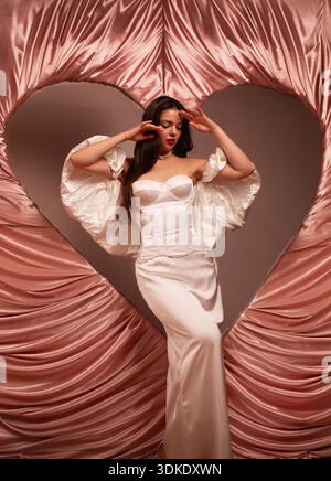 Una donna indossa con eleganza un abito bianco sullo sfondo rosa a forma di cuore mentre celebra l'amore a San Valentino. Foto Stock