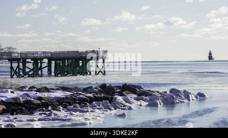 NORWALK, CT, USA - 30 GENNAIO 2026: Un Long Island Sound ghiacciato si estende sotto un cielo limpido. Un molo di legno si estende nell'acqua, mentre un faro Foto Stock