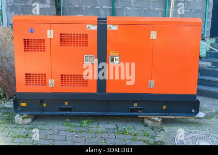 Generatore industriale per esterni con involucro arancione su blocchi di cemento vicino alla parete in mattoni Foto Stock