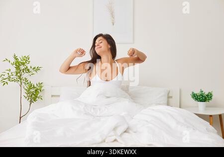 Ritratto di una giovane donna felice sorridente che si sveglia a letto allungando le braccia nella camera da letto moderna. Foto Stock
