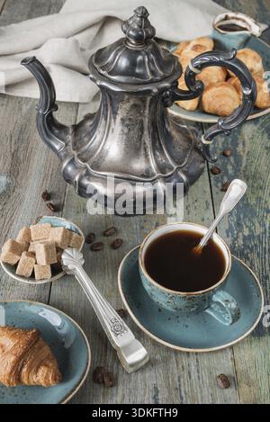 Caffè nero in tazza vintage, croissant su un vecchio piatto blu e una bellissima caffettiera d'argento su vecchie tavole di legno scuro Foto Stock