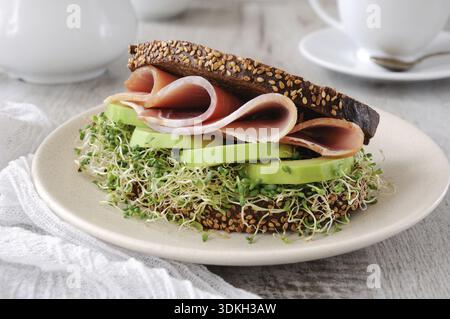 Cibo, panino di pane di segale con cereali, fette di prosciutto e avocado con germogli di erba medica germogliata Foto Stock