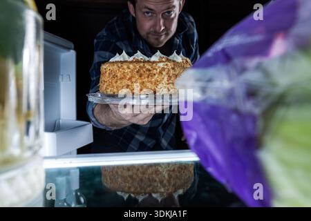Un creativo punto di vista (POV) dall'interno di un frigorifero illuminato. L'immagine cattura l'espressione gioiosa di un uomo mentre lui Foto Stock