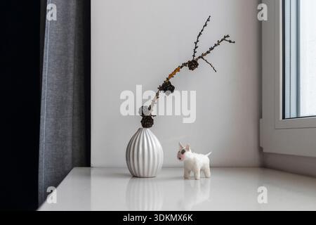Composizione interna scandinava con vaso in ceramica scanalata bianca che contiene rami secchi con coni di pino, piccola statuetta bianca di animali (toro o mucca), e. Foto Stock