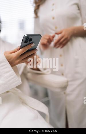Primo piano ritagliato di donna e cosmetologa impegnata in consulenza, cliente che tiene lo smartphone per fare riferimento a informazioni visive sull'estetica Foto Stock