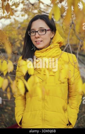 Bella donna sorridente con giacca gialla e sciarpa sullo sfondo di una foresta autunnale Foto Stock