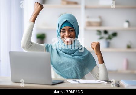 Grandi notizie. Felice entusiasta Black Muslim Lady che celebra il successo con il portatile Foto Stock