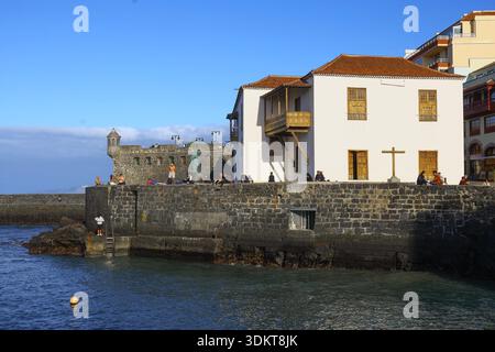 Puerto de la Cruz, Tenerife, 01152026 - edificio bianco sul mare con persiane in legno e balcone situato su un molo di pietra, con persone che camminano lungo il mare Foto Stock