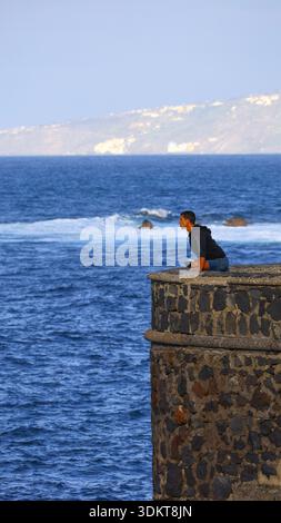 Puerto de la Cruz, Tenerife, 01152026 — Un corridore accovacciato su una sporgenza di pietra sopra l'oceano, che guarda le onde e la costa lontana in una diga Foto Stock