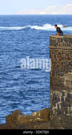 Puerto de la Cruz, Tenerife, 01152026 — Un corridore che si estende su una struttura in pietra che si affaccia sull'oceano, inclinato in avanti dopo una corsa mentre osserva il Foto Stock