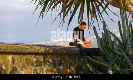 Puerto de la Cruz, Tenerife, 01152026 — un uomo anziano seduto su un muro di pietra circondato da vegetazione tropicale, tra cui cactus e piante simili a palme Foto Stock
