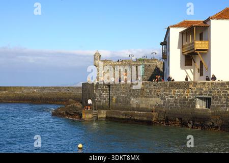 Puerto de la Cruz, Tenerife, 01152026 - molo di pietra che si estende nelle calme acque costiere con persone che camminano lungo il lungomare, incorniciato da uno storico lungomare Foto Stock