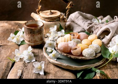 Piatto in ceramica con dolci mochi colorati teiera da tè e fiori di gelsomino bianchi su un rustico tavolo in legno con panno di lino Foto Stock