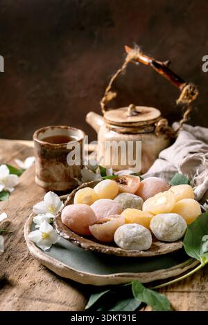 Colorate torte di riso mochi su piatto di ceramica con tradizionali fiori di gelsomino teiera su un rustico tavolo di legno che crea un autentico bancomat giapponese per la cerimonia del tè Foto Stock