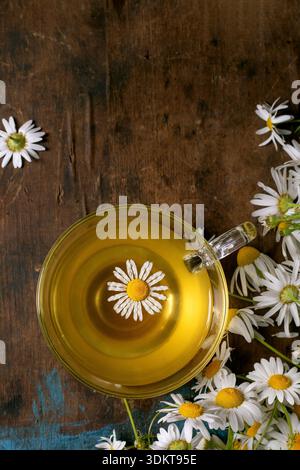 Tazza di tè di camomilla in vetro con fiori di margherita galleggianti circondati da fiori di camomilla freschi su uno sfondo rustico in legno. Concetto di tè alle erbe con remed naturale Foto Stock