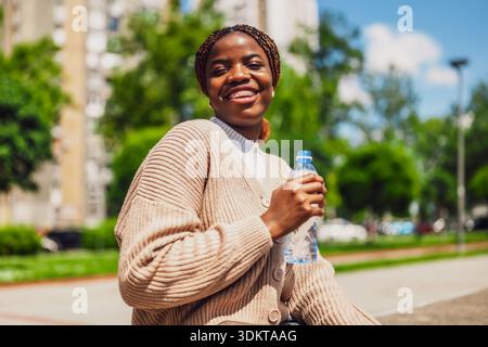 Ritratto di una giovane donna nera allegra che si gode una giornata di sole in città. Sta tenendo una bottiglia d'acqua. Foto Stock