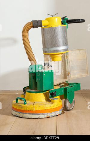Una lucidatrice per pavimenti verde e giallo con un'unità antipolvere cilindrica e un tubo flessibile si trova su un pavimento in legno usurato contro una parete liscia di colore chiaro. Foto Stock