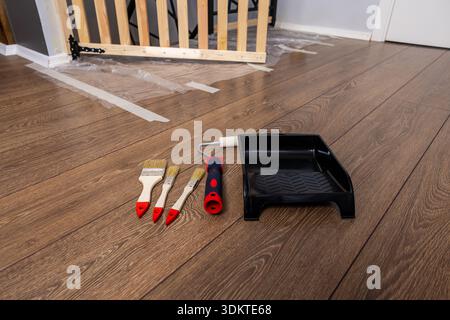 Set di pennelli, un rullo per pittura e un vassoio nero su un pavimento in legno con portabicchieri parzialmente assemblato e materiali di ristrutturazione nel backgroun Foto Stock