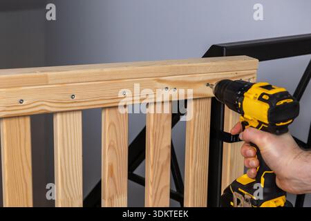 Una persona fissa le viti in una struttura in legno naturale con un trapano elettrico giallo e nero. Sullo sfondo è visibile una cornice in metallo nero. Foto Stock