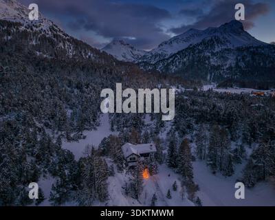 Vista aerea dei pini innevati che abbracciano una cabina solitaria mentre il crepuscolo dipinge il cielo su maestose montagne innevate, Maloja, Grigioni, Svizzera. Foto Stock