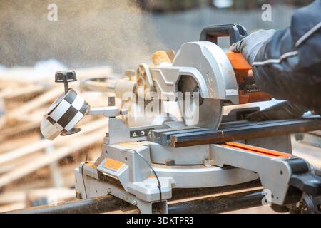 Primo piano di una sega circolare in uso, tagliando il legno con la segatura che vola. Un guanto guida l'utensile, con tavole di legno e una recinzione sullo sfondo. Foto Stock