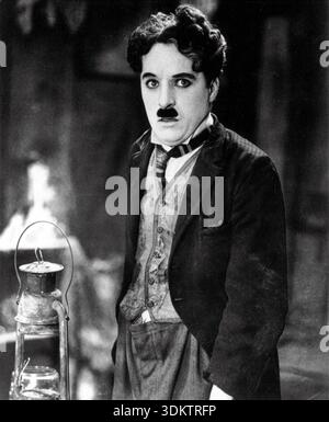 Questa iconica fotografia in bianco e nero degli anni '1920 mostra Charlie Chaplin come il suo amato personaggio, The Tramp, in una scena della "corsa all'oro". Si erge con un'espressione leggermente preoccupata, vestito con i suoi abiti sovradimensionati riconoscibili e i suoi piccoli baffi, accanto a una vecchia lanterna con quello che sembra essere vapore o fumo sullo sfondo. Foto Stock