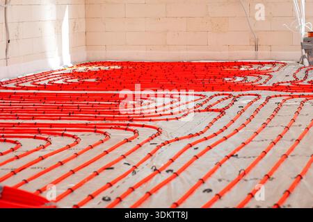 Interno di un cantiere che mostra tubi di riscaldamento rossi a pavimento disposti in anse su uno strato di base, con pareti in mattoni chiari e componenti elettrici visibili Foto Stock