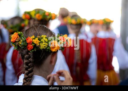 Un gruppo di bambini vestiti con costumi nazionali lettoni con strisce rosse e bianche. Corone floreali di fiori arancioni e gialli adornano le loro teste. Foto Stock