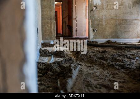 Un cantiere interno che mostra pareti non finite, cemento bagnato con scanalature, tubi esposti e un corridoio poco illuminato con una scala in vista. Foto Stock