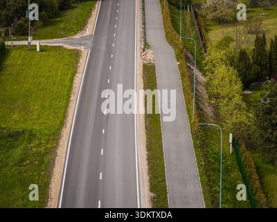 Una strada dritta a due corsie con segnaletica bianca corre lungo un sentiero pedonale, delimitato da erba, alberi, recinzione e semafori. Foto Stock