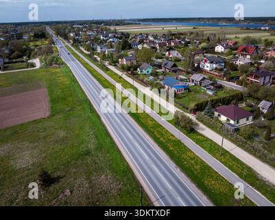 Una strada dritta a due corsie corre tra i campi e un'area residenziale con case colorate, giardini e alberi. Una foresta e l'acqua sono visibili nel di Foto Stock