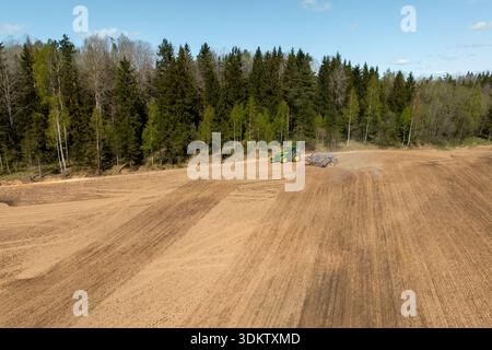 Un trattore verde lavora vicino a un cumulo di materiale in un campo coltivato con motivi di terreno visibili, delimitato da alberi sempreverdi e decidui sotto una b limpida Foto Stock