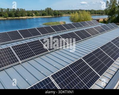 I pannelli solari su un tetto metallico si affacciano sul sole, con un corpo d'acqua delimitato da alberi e vegetazione visibile in lontananza. Foto Stock