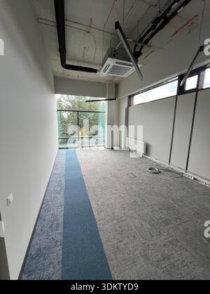 Spazio ufficio vuoto con soffitto in cemento, tubi esposti, grandi finestre, tappetini blu e grigi, radiatore e cablaggio elettrico visibile lungo le pareti. Foto Stock