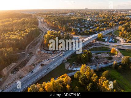 La vista aerea mostra lo svincolo autostradale su più livelli in fase di costruzione, con un cavalcavia sopraelevato, macchine per il movimento terra, pile e folia mista verde e ambra Foto Stock