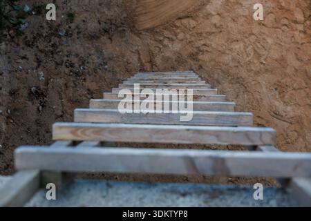 Vista dall'alto verso il basso di una scala di legno che conduce a un profondo sito di scavo, mostrando strati di terreno compattati, sporcizia e detriti sparsi intorno alla fossa. Foto Stock