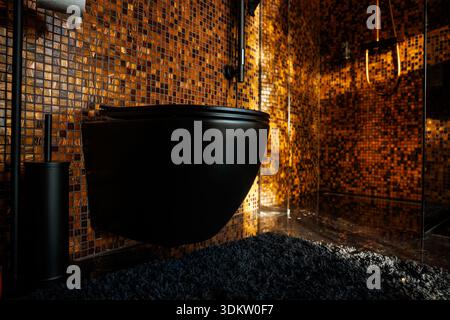 Bagno dal design contemporaneo con WC a parete nero, pareti piastrellate a mosaico in arancione e marrone, divisorio doccia in vetro e tappeto scuro testurizzato. Foto Stock