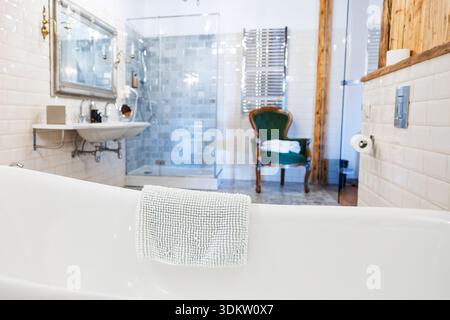 Una vasca da bagno bianca e un box doccia in vetro con piastrelle a motivi geometrici si trovano vicino ai radiatori cromati. Una poltrona in velluto verde e uno specchio in stile antico incorniciano un floati Foto Stock