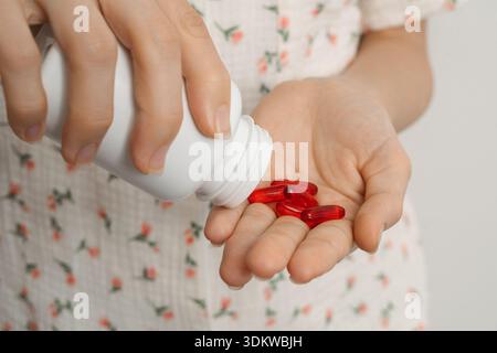 Primo piano di una donna in pigiama floreale che tiene in mano una bottiglia bianca e le versa capsule rosse. Concetto di assistenza sanitaria, farmaci, vitamine, e. Foto Stock