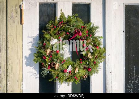 Tradizionale corona natalizia fatta di rami sempreverdi, coni di pino e bacche rosse appese su una porta rustica di legno. Corona natalizia fatta a mano. Foto Stock