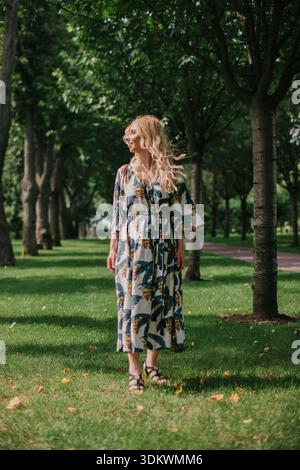 Donna che cammina in un parco verde con maxi abito floreale, vento che soffia attraverso i capelli Foto Stock
