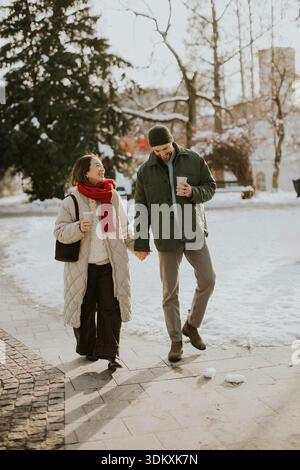 Una donna coreana e un uomo caucasico camminano mano nella mano attraverso un parco coperto di neve. Entrambi stanno tenendo le tazze e sembrano godersi una giornata invernale Foto Stock
