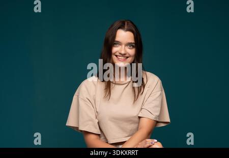 Giovane donna sorridente con trucco naturale e capelli marroni in posa su sfondo verde scuro in studio con espressione rilassata e amichevole Foto Stock