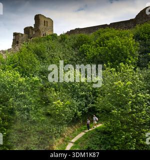 Una coppia che cammina su un sentiero sotto le mura di pietra del Castello di Scarborough, Yorkshire, Regno Unito. La fortezza sorge su un promontorio che domina la città. Foto Stock