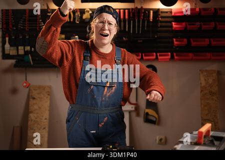 La donna falegname indossa indumenti da lavoro in officina con attrezzi appesi alla parete che scuote il pugno fissato alla telecamera minacciando. falegname donna in tarda serata Foto Stock