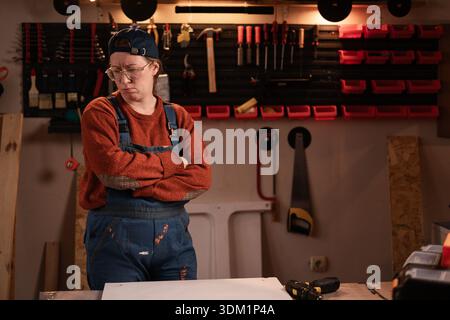 La donna falegname indossa indumenti da lavoro sul posto dell'officina con attrezzi appesi al risentimento delle pareti in caso di conflitto o disaccordo. falegname femminile eveni tardivi Foto Stock