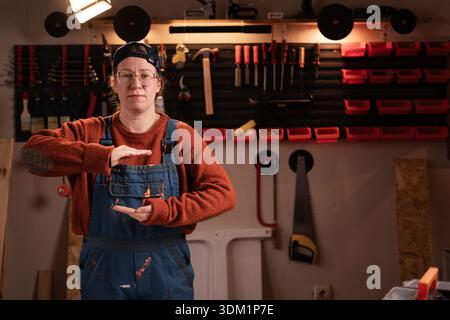La donna falegname indossa indumenti da lavoro in officina con attrezzi appesi alla parete che mostrano una grande scatola prodotto di grandi dimensioni con gesti delle mani. falegname femminile eveni tardivi Foto Stock