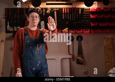 La donna falegname indossa indumenti da lavoro in officina con attrezzi appesi alla parete che fanno fermare il movimento guardando la fotocamera. falegname donna in tarda serata Foto Stock